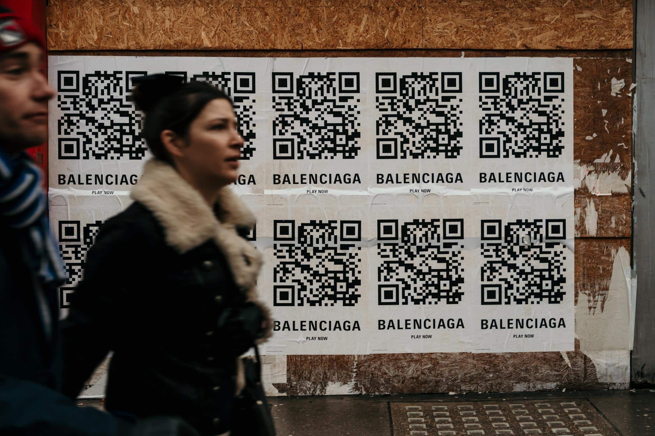 Modelabel Balenciaga nutzt QR-Codes in Größe A2 als Werbung. Plakate hängen an Holzwand und Frau läuft vorbei.