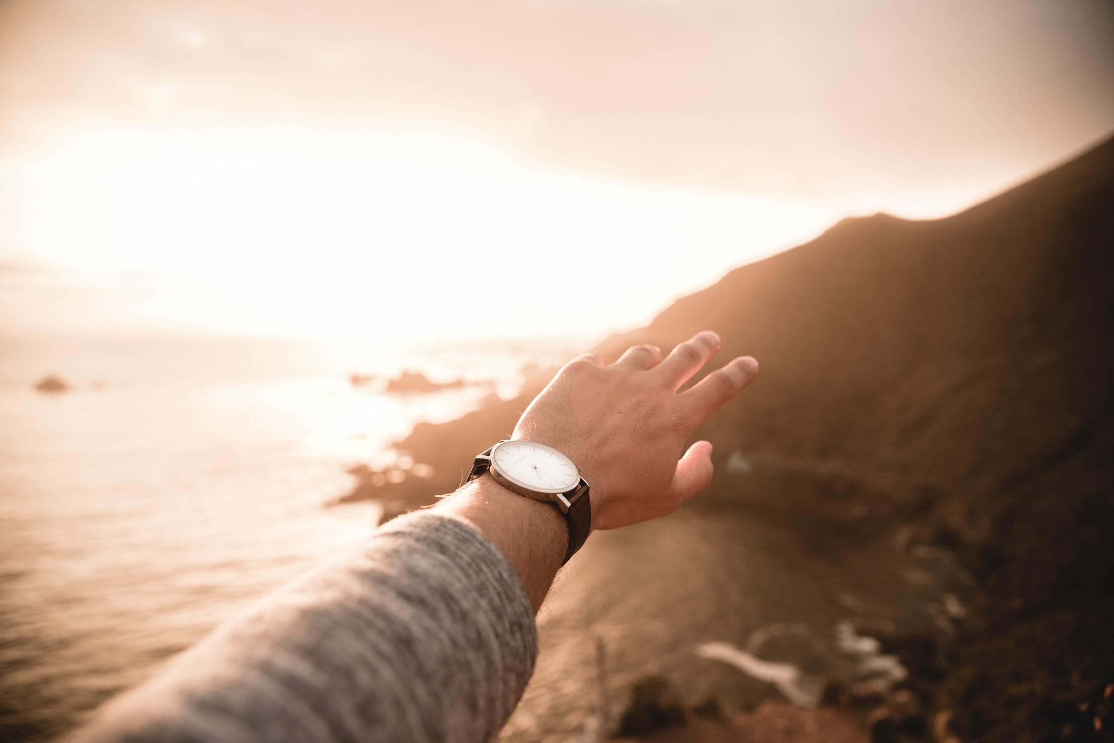 Ausgestreckte Hand vor Meer und Hügeln. Die Hand hat eine Armbanduhr am Handgelenk.