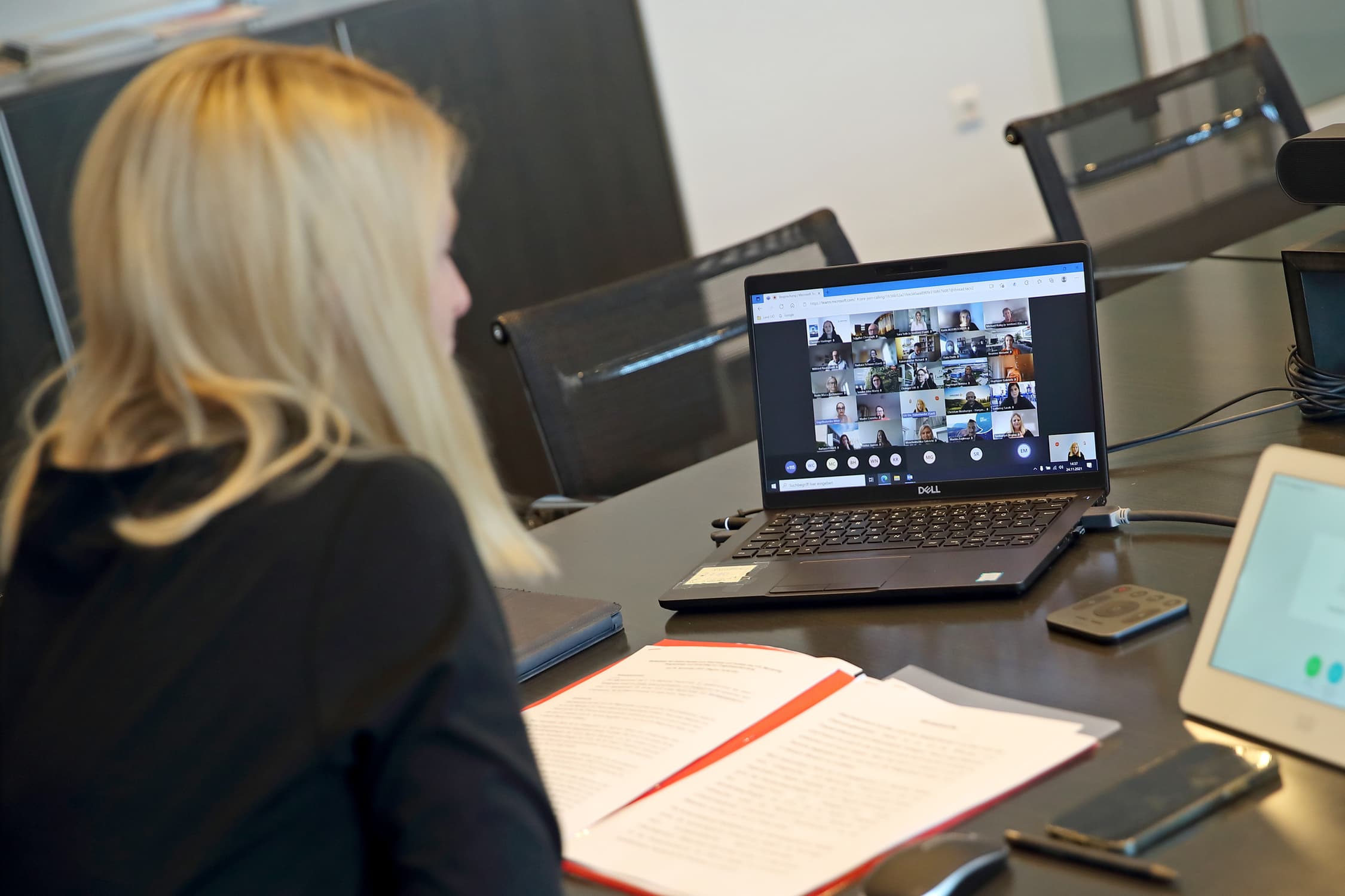 Aufnahme eines Bildschirms, auf dem man einen Videocall mit mehreren Menschen sieht. Eine blonde Frau sitzt vor dem Laptop mit dem Rücken zur Kamera.