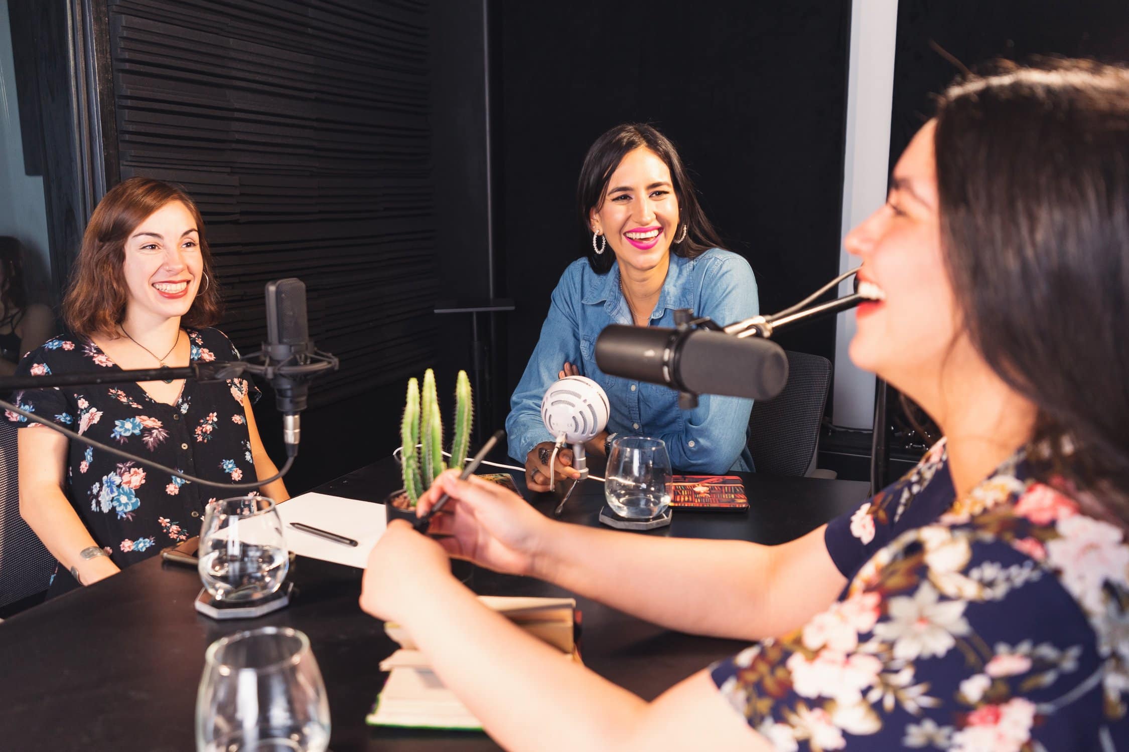3 junge Frauen sitzen am Tisch und unterhalten sich. Eine von ihnen spricht wie in einem Podcast-Setting in ein Mikrofon.