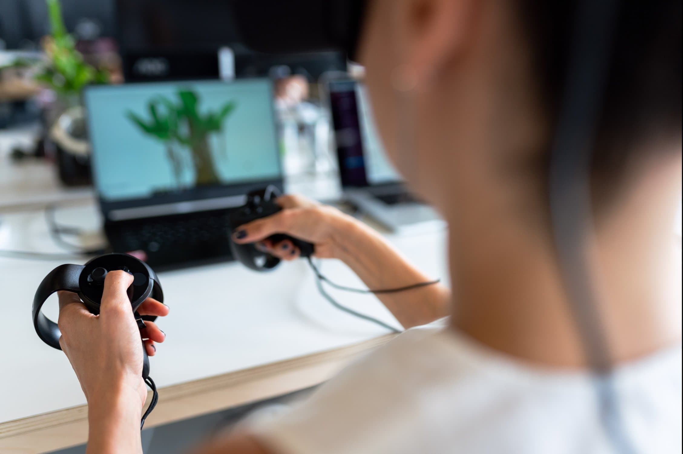 junge Frau von hinten, die mit Controllern in der Hand vor einem Schreibtisch am Laptop sitzt.