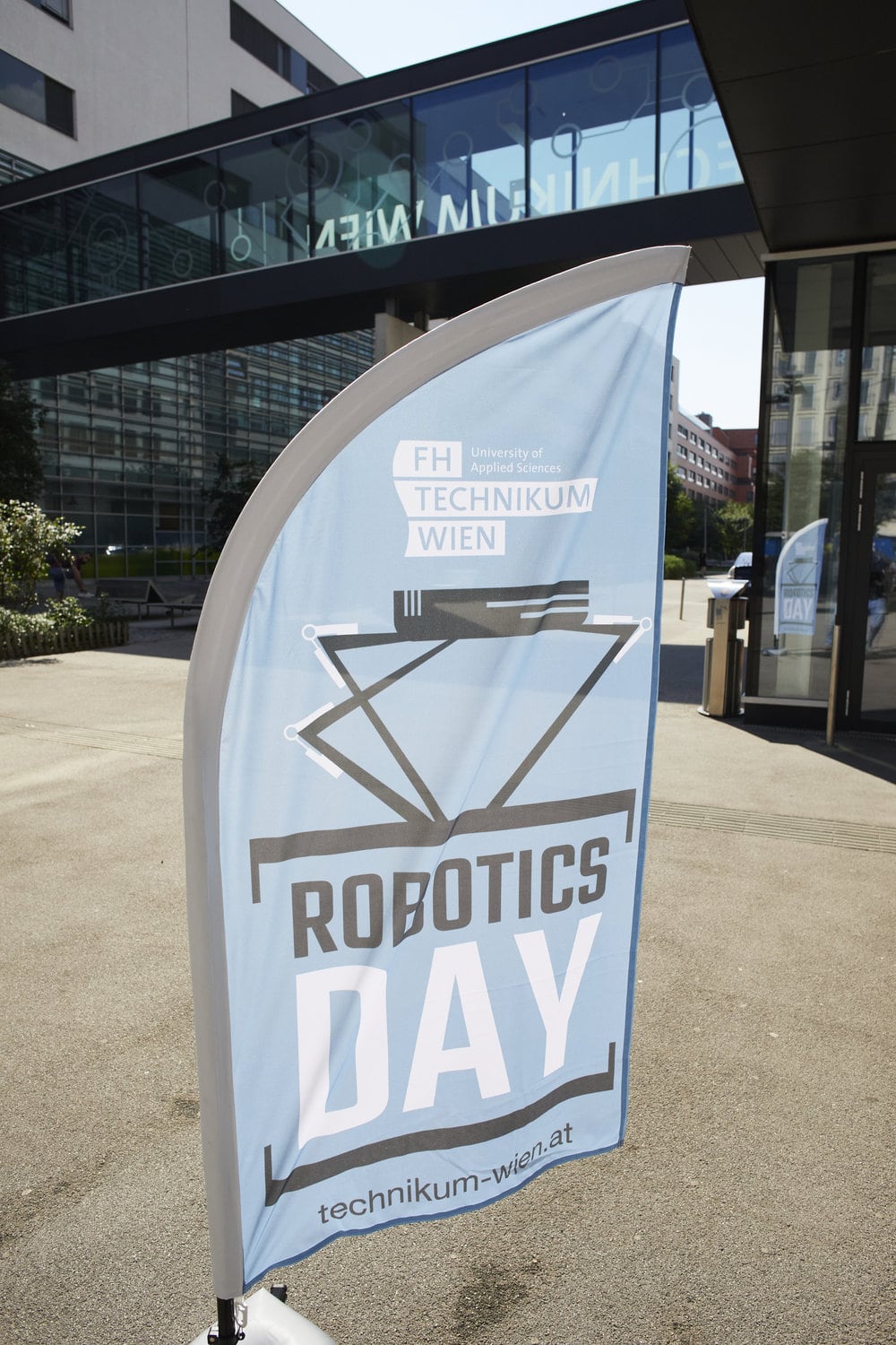 Eine blaue Beachflag mit der Aufschrift Robotics Day 2023 vor einem Campus-Hintergrund.