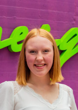 Junge Frau mit roten Haaren steht vor einer lila Wand mit grüner Schrift darauf.