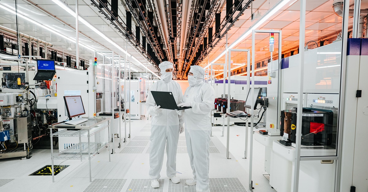 Reinraum bei Infineon. Zwei Menschen stehen in weißer Schutzkleidung mit einem Laptop in der Hand in einem Labor.
