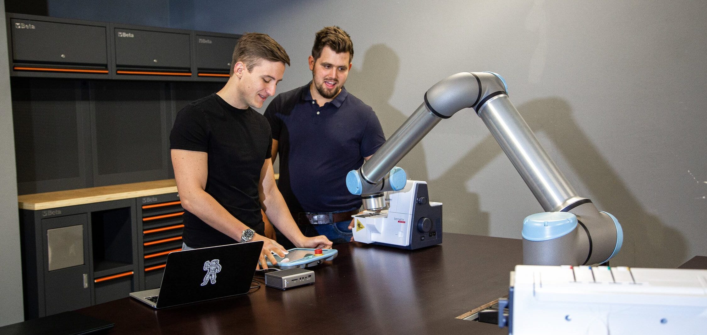 Zwei junge Männer bedienen einen Roboterarm.