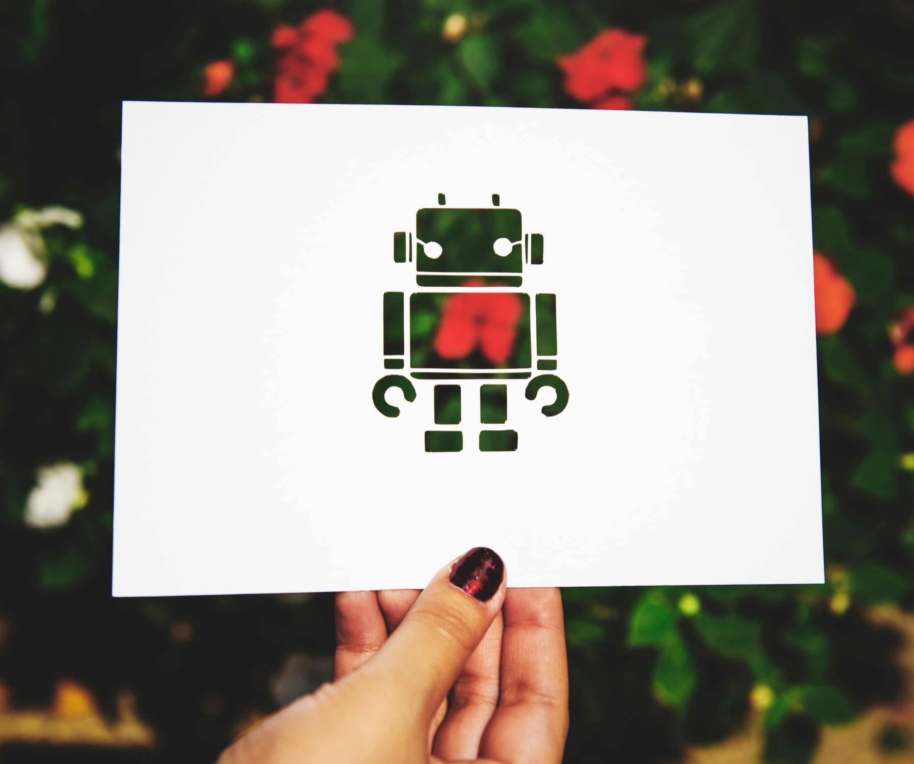 Eine Hand hält ein Papier vor einen Hintergrund mit Blumen, auf dem ein Roboter ausgeschnitten wurde.