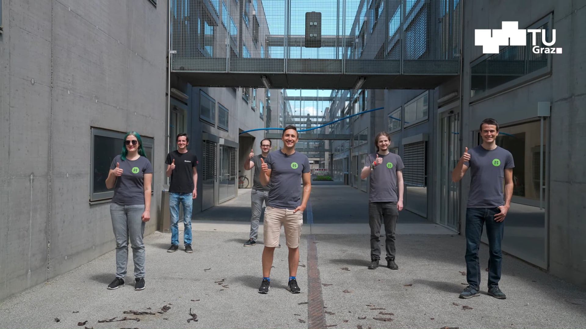 LosFuzzys der TU Graz stehen in aspahltiertem Innenhof und zeigen mit dem Daumen nach oben.