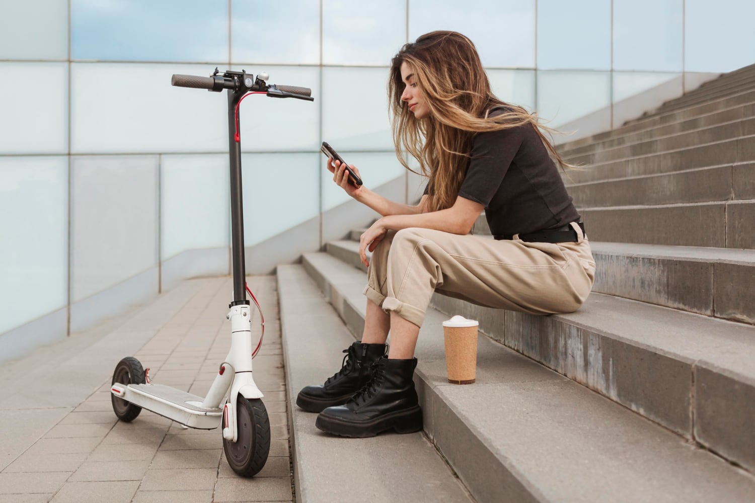 Junge Frau sitzt auf einer Treppe im freien. Sie schaut auf ihr Handy. Neben ihr stehen ihr E-Roller und ein Coffe-to-go-Becher.