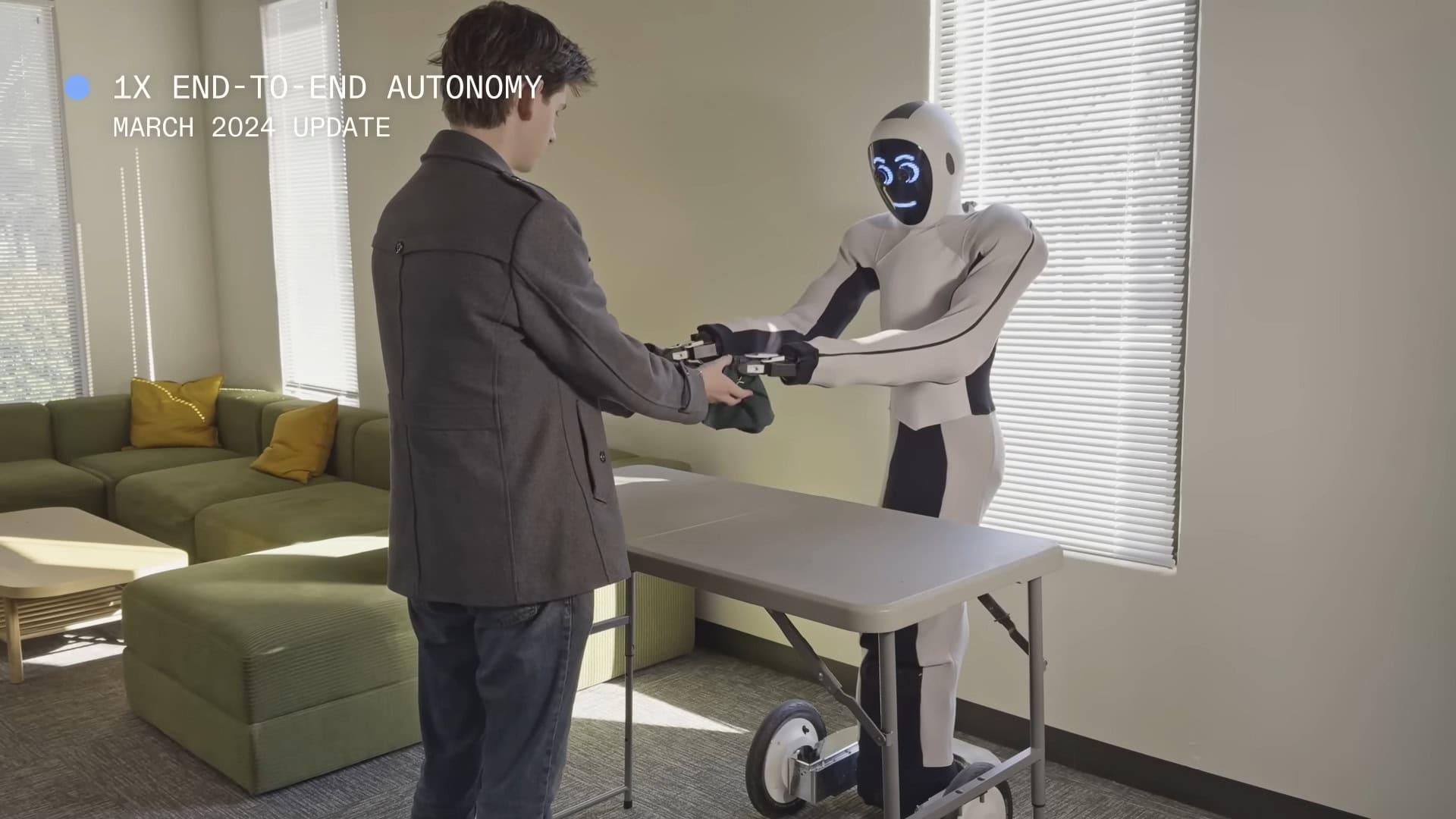 Menschengroßer Roboter steht hinter Tisch und hält gegenüberliegenden Mann an den Händen. Das ganze findet in einem Raum statt, der Ähnlichkeit mit einem Wohnzimmer hat.