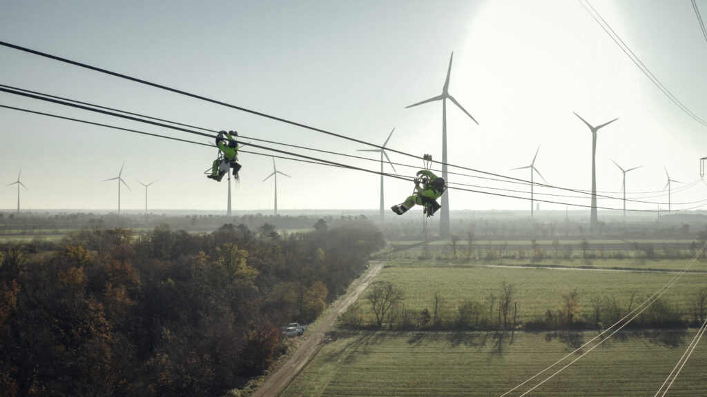 Zwei Personen hängen mit Gurten an Freileitungen. Im Hintergrund sieht man ein paar Windräder.