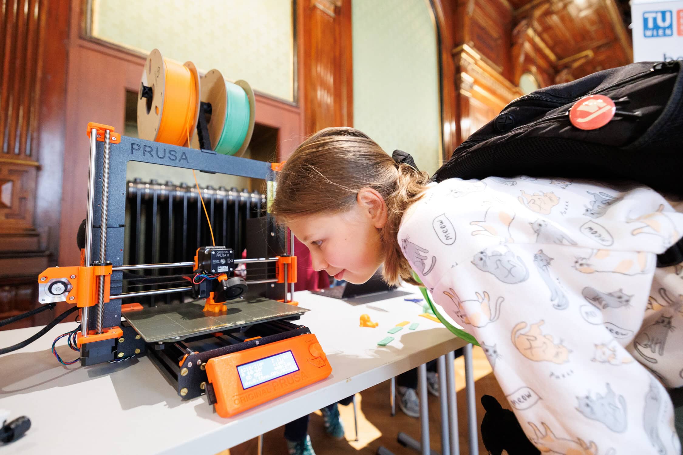 Ein Mädchen mit einem weißen Pullover und einem schwarzen Rucksack am Rücken schaut interessiert dabei zu, was eine auf einem Tisch stehende Maschine gerade macht.