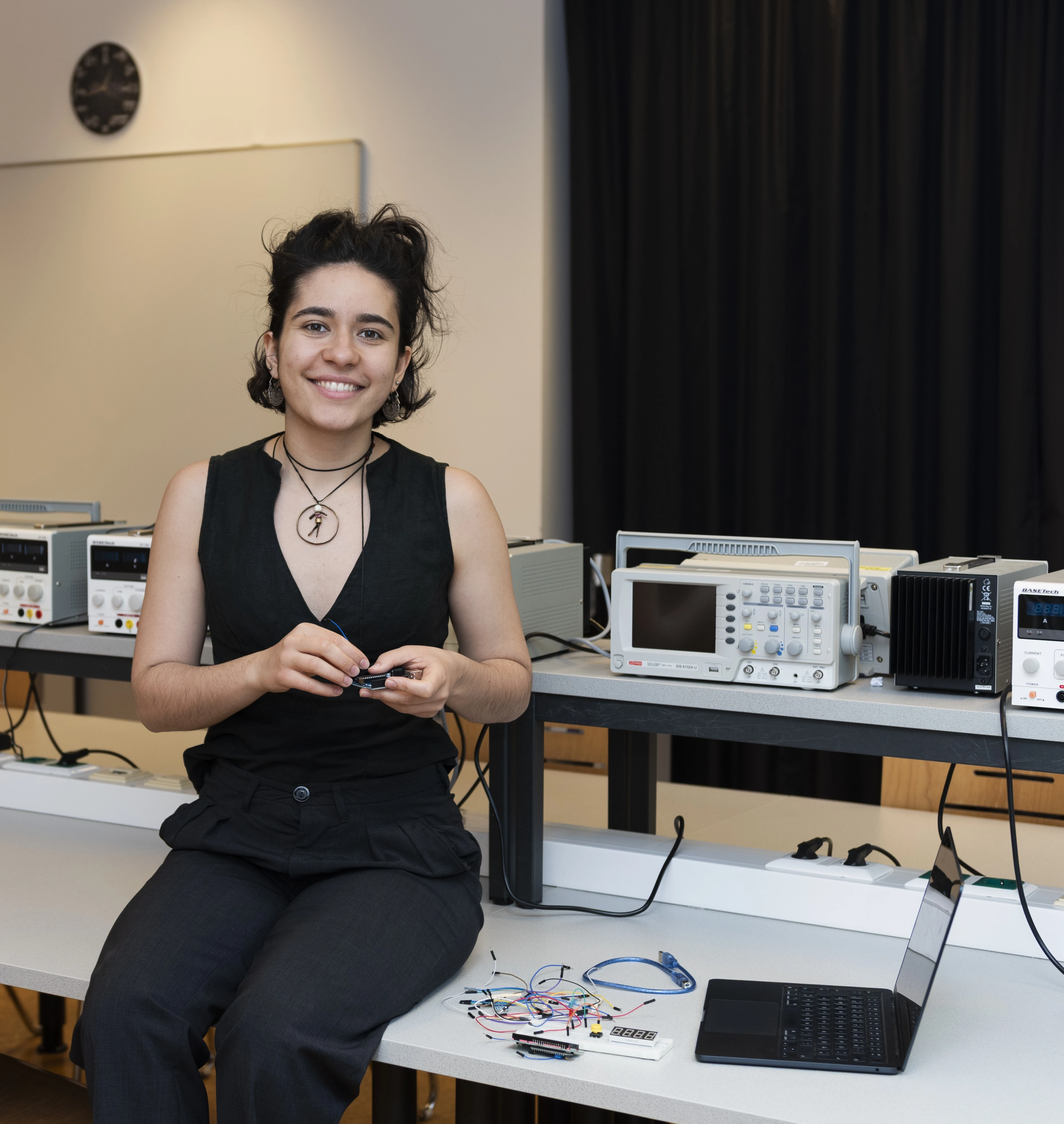 Volt&Byte-Workshopleiterin Sonja Schobesberger sitzt auf einem Schreibtisch, auf dem ein Laptop und ein paar Kabel liegen. Im Hintergrund sind ein paar technische Geräte auf Schreibtischen zu sehen.