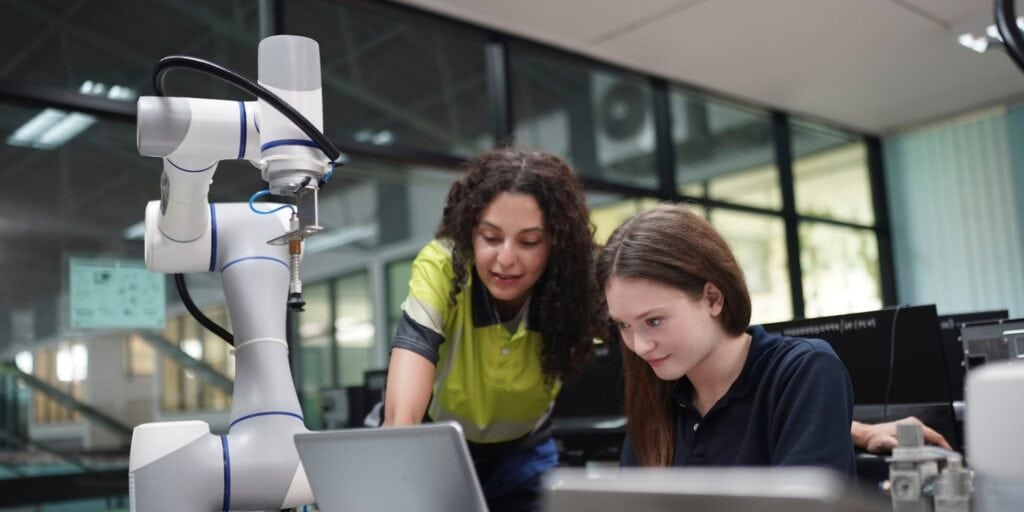 Zwei junge Frauen arbeiten neben einem Roboterarm am Laptop.