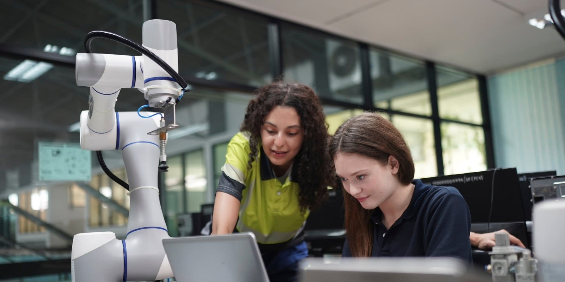 Zwei junge Frauen arbeiten neben einem Roboterarm am Laptop.