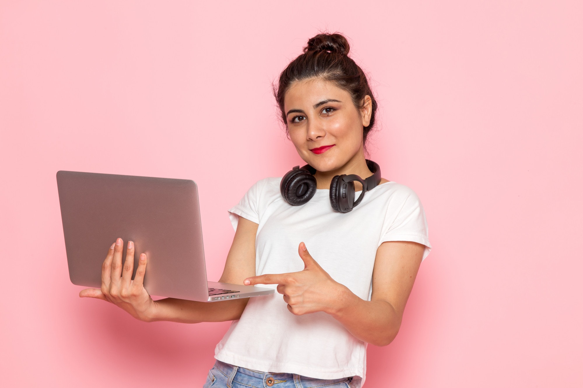 Eine Jugendliche mit hochgeundenen Haaren und rot geschminkten Lippen steht vor einem rosa Hintergrund. Sie trägt Kopfhörer um den Hals unnd hat einen Laptop in einer Hand, auf den sie mit der anderen Hand deutet.