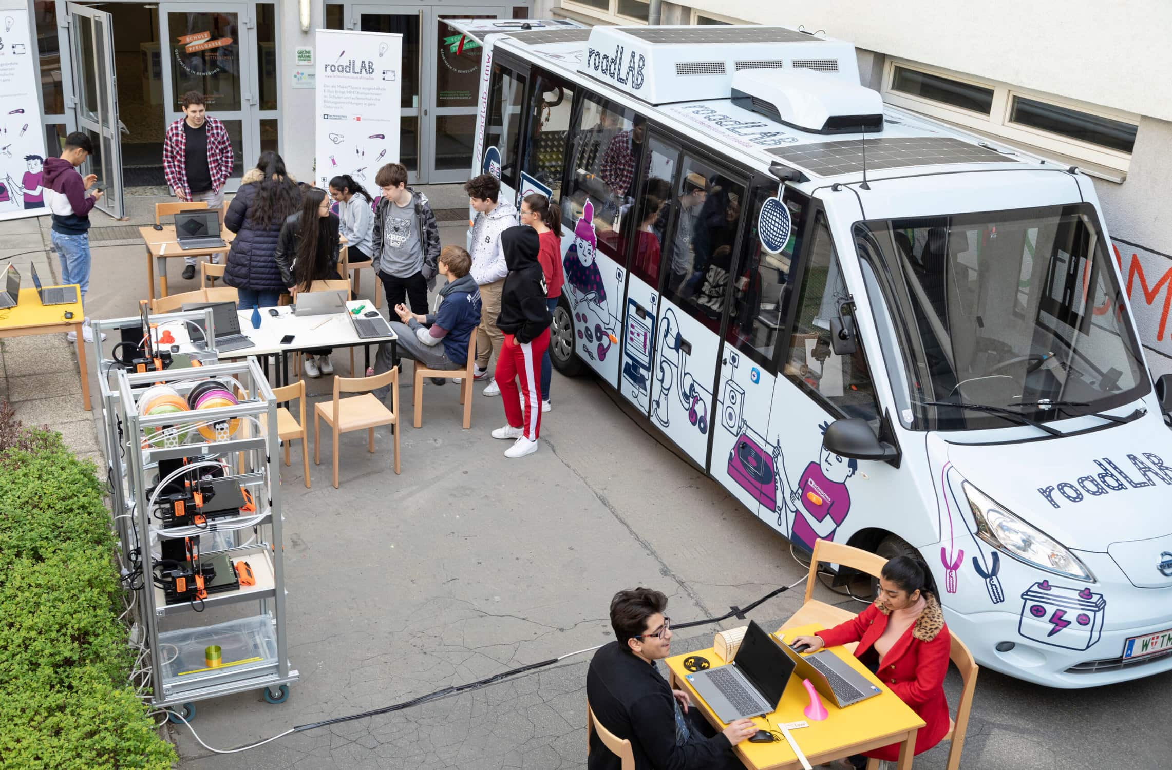 Das RoadLAB steht auf einem Parkplatz, daneben sind Tische und Stühle aufgebaut, auf denen Jugendliche sitzen und technisch-kreativ arbeiten