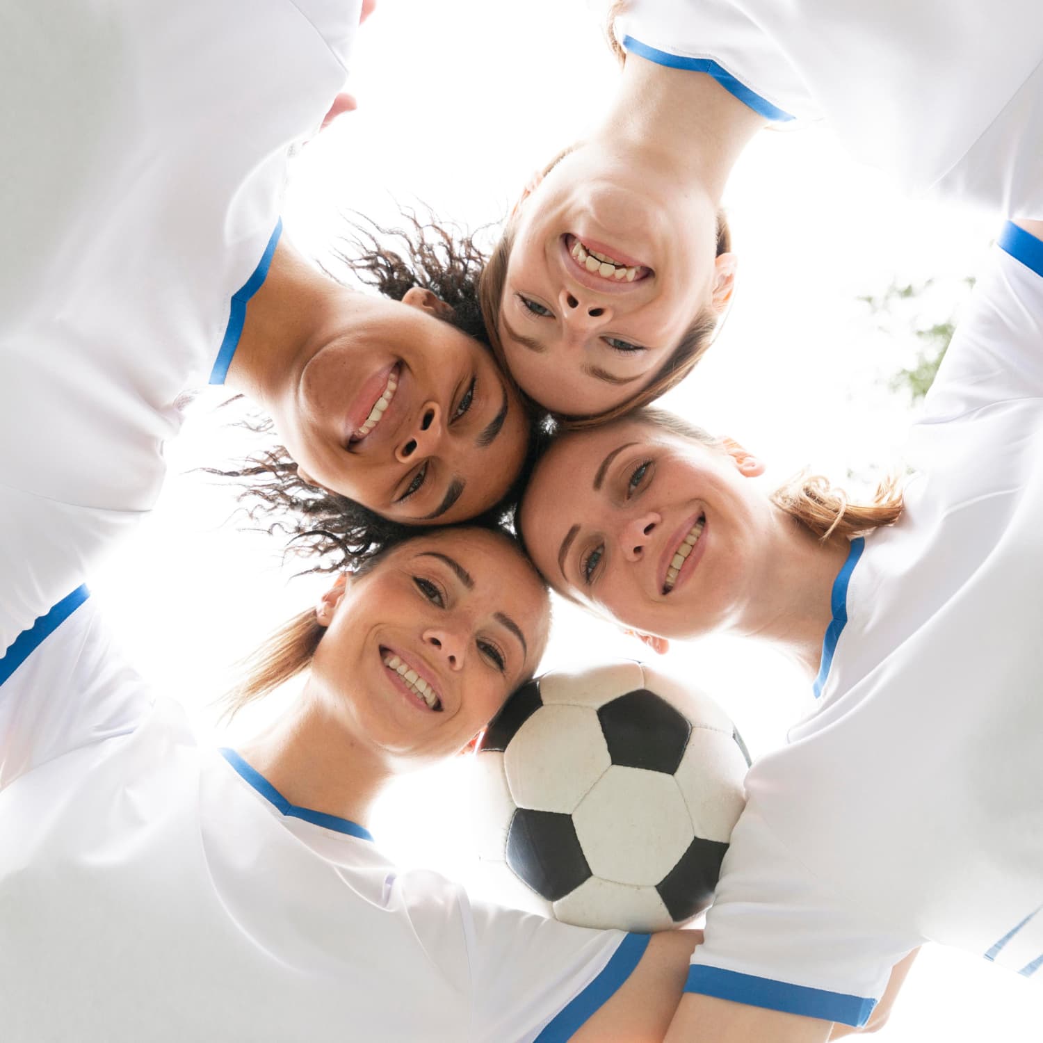 Vier lächelnde junge Frauen in heller Sportkleidung, die die Köpfe im Kreis mit einem Fußball auf den Schultern aneinander halten