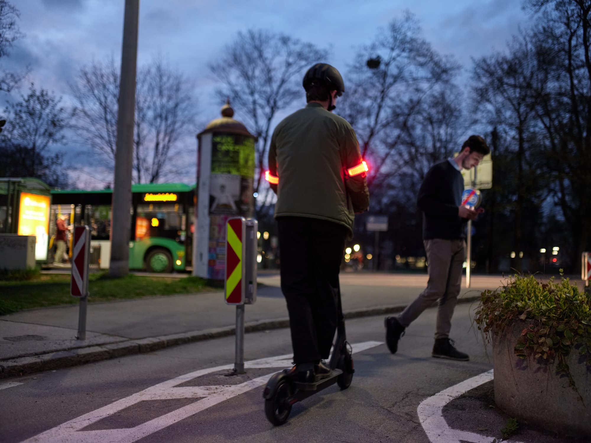 E-Scooter-Fahrer am Abend, mit zwei rot leuchtenden Armbändern von Flasher an den den Oberarmen.